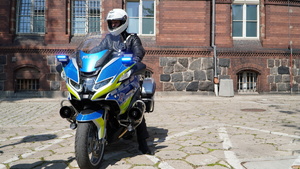 Zdjęcie przedstawia policjanta na motocyklu przed budynkiem KMP w Olsztynie.