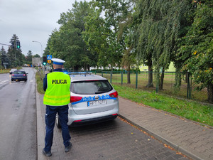 Zdjęcie przedstawia policjanta oraz radiowóz, a w tle szkoła i przejście dla pieszych.