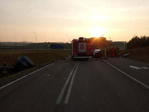 Ilustracja przedstawia miejsce wypadku drogowego. Na zdjęciu widoczne uszkodzone pojazd osobowe oraz funkcjonariusze służb ratunkowych odpowiedzialnych za bezpieczeństwo.