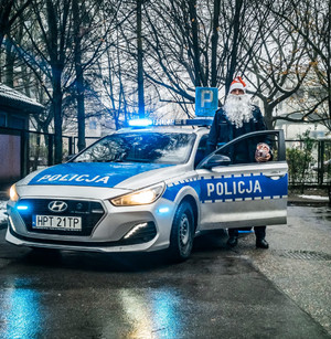 fot.st.sierż. Jarosław Nożyński . Zdjęcie przedstawia funkcjonariusza w czapce Mikołaja i białej brodzie na tle radiowozu.