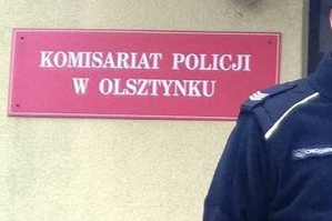 Zdjęcie przedstawia fragment policjanta, a w tle budynek Komisariatu Policji w Olsztynku.