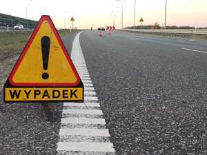 Zdjęcie przedstawia znak drogowy "Wypadek" oraz w tle policjantów oraz auto na drodze szybkiego ruchu.