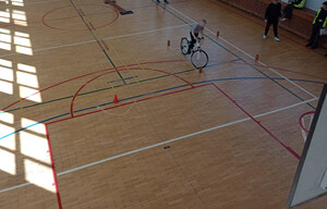 Zdjęcie przedstawia uczestników turnieju na hali sportowej. W centrum znajduje się chłopiec pokonujący tor przeszkód.