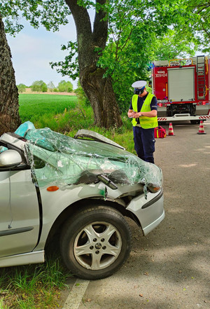 Zdjęcie przedstawia miejsce wypadku drogowego, pracujące służby oraz auto, które uderzyło w drzewo.