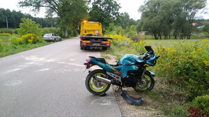 Zdjęcie przedstawia miejsce zdarzenia drogowego, motocykl oraz lawetę, w tle pojazd osoby na drodze.