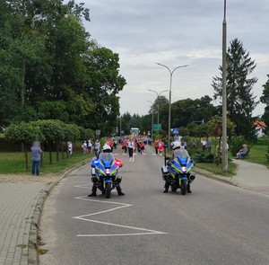 Zdjęcie przedstawia policyjnych motocyklistów na motorach oraz w tle przemarsz.