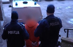Zdjęcie przedstawia zatrzymanego mężczyznę prowadzonego przez umundurowanych policjantów.
